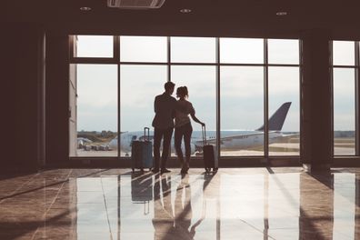 Rear view of couple standing in waiting room in the airport departure area in front of window with airplane in the background, wide angle view  at sunset.