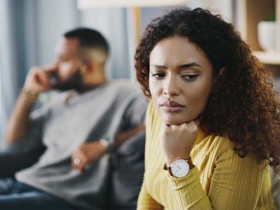 A man and woman sitting on the couch. The woman is in the foreground looking upset.