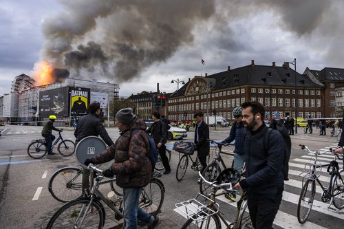 People ride bicycles as smoke rises from the Old Stock Exchange in Copenhagen, Denmark, Tuesday, April 16, 2024.